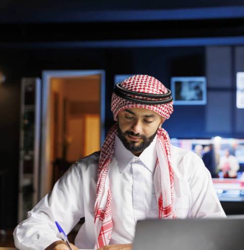 Professional Muslim guy uses technology to work efficiently in the office, browsing online and writing notes from a laptop. Youthful Arab male student using his notebook and minicomputer for research.